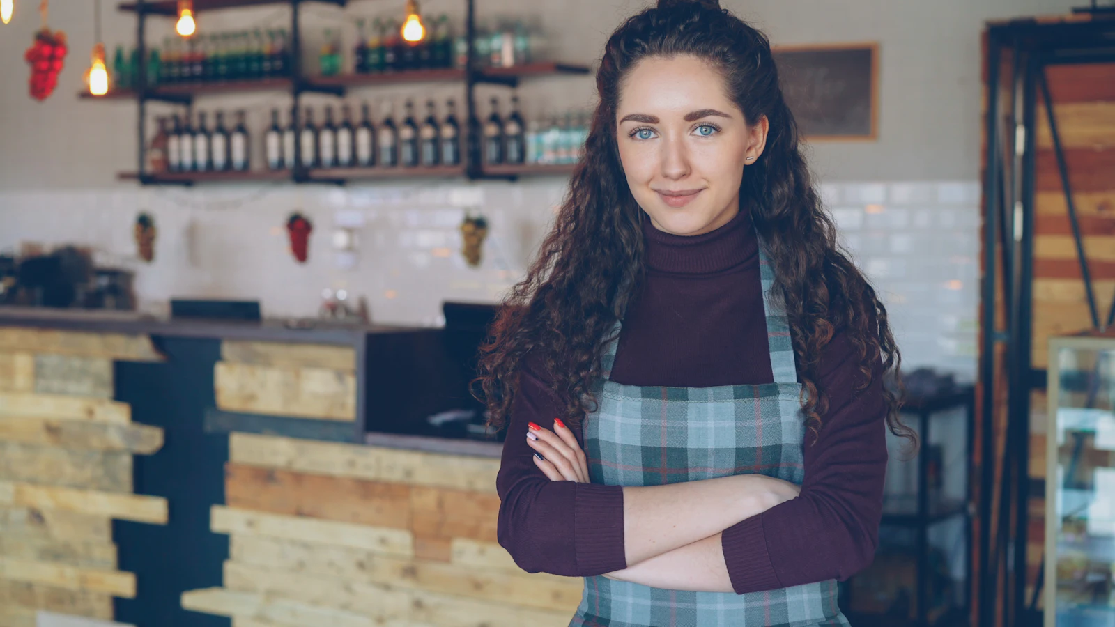 Eine Frau mit Schürze steht mit verschränkten Armen in einem kleinen Café vor der Theke